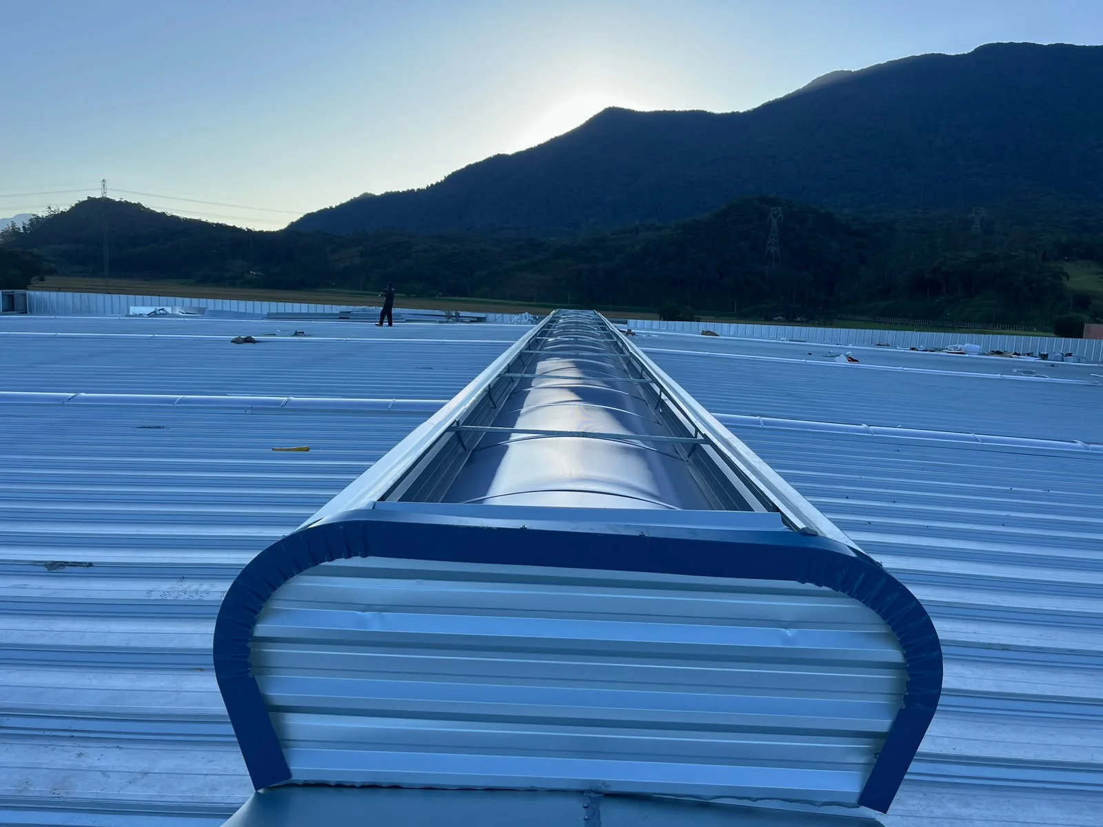 Cobertura de galpão com lanternim de iluminação natural ao amanhecer, com montanhas ao fundo